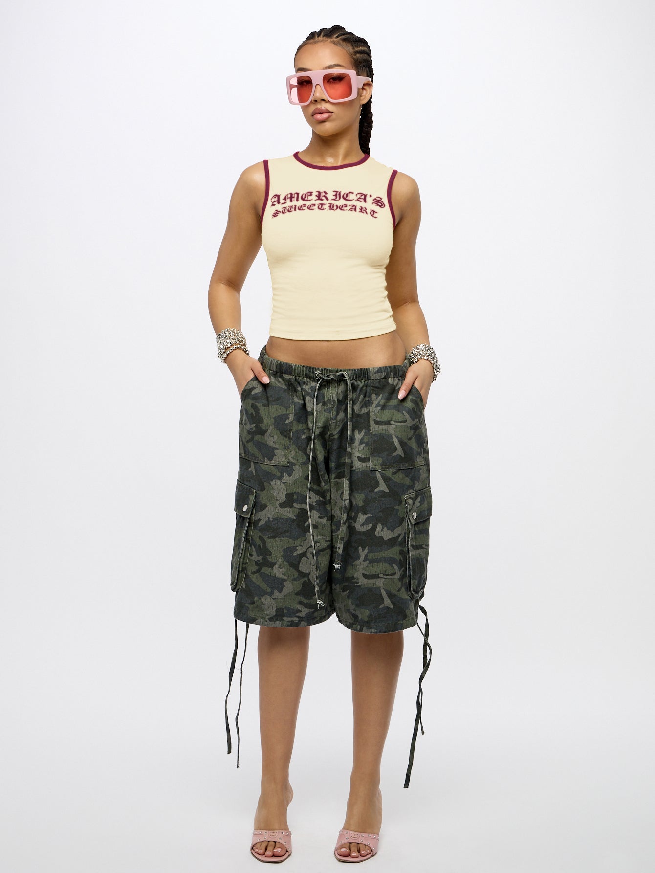 A model stands against a light gray studio background wearing a cream ribbed crop top with red trim and text, green camouflage cargo shorts with side laces, pink sunglasses, bracelets, and pink heeled sandals.