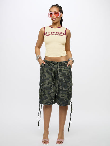 A model stands against a light gray studio background wearing a cream ribbed crop top with red trim and text, green camouflage cargo shorts with side laces, pink sunglasses, bracelets, and pink heeled sandals.